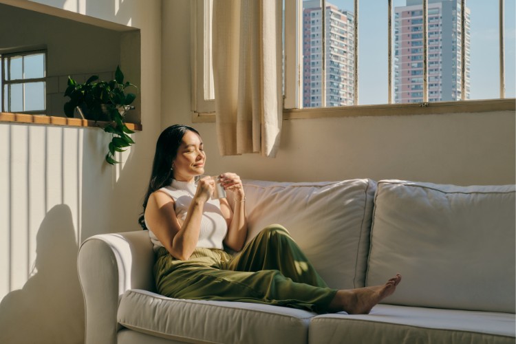 A woman sits on a white sofa in a sunlit room, holding a cup and smiling peacefully. She wears a white shirt and green pants. Large windows reveal a view of tall buildings outside. A plant sits on a ledge in the background.