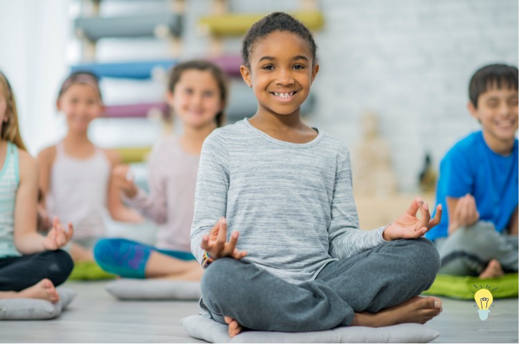 A smiling child sitting cross-legged on a cushion, with hands in a meditation pose. Other children in the background are also meditating. The setting appears to be a yoga or meditation class.