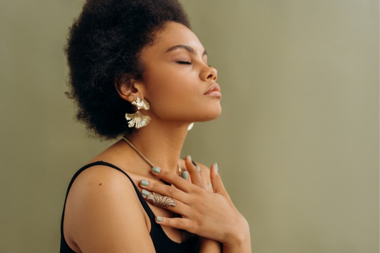 A person with closed eyes stands against a light green background. They have short curly hair, wear a black sleeveless top, large feather-shaped earrings, and a necklace. Their hands, adorned with rings, are gently placed on their chest.