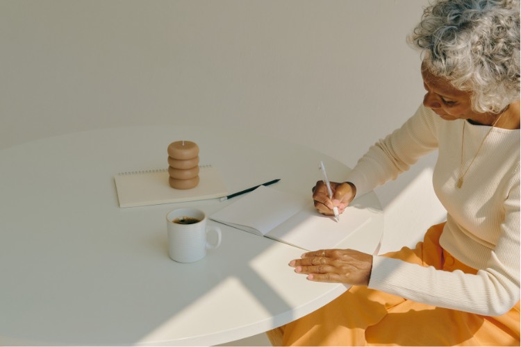 An elderly person with grey hair sits at a round white table, writing in a notebook. A cup of coffee, a pen, a stacked candle, and another notebook are on the table. Sunlight casts soft shadows, creating a serene atmosphere.