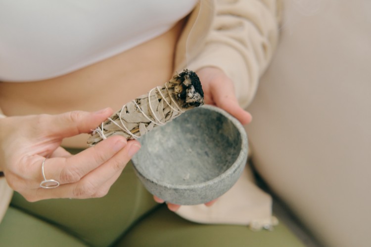 A person holding a bundle of burning sage wrapped with string in one hand and a small stone bowl in the other. The person is seated, wearing a beige top and green pants. The background is softly blurred.